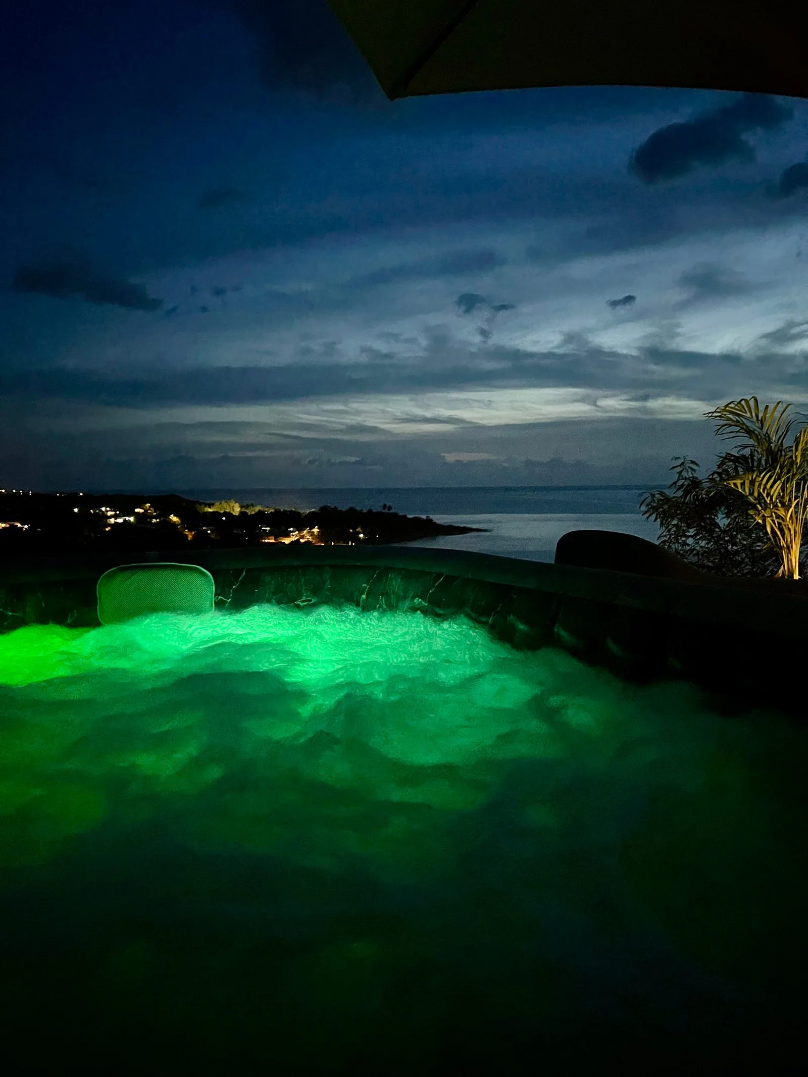 Jacuzzi illuminé en vert néon la nuit avec vue sur la mer et les lumières de la côte